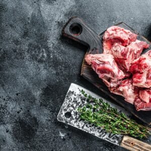 Raw lamb meat stew cuts with bone on wooden butcher board and cleaver. Black background.