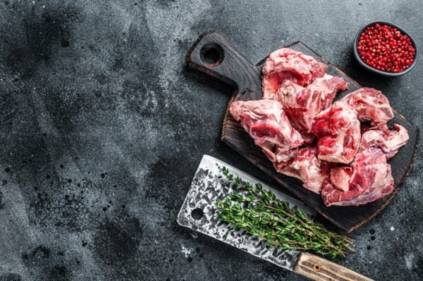 Raw lamb meat stew cuts with bone on wooden butcher board and cleaver. Black background.